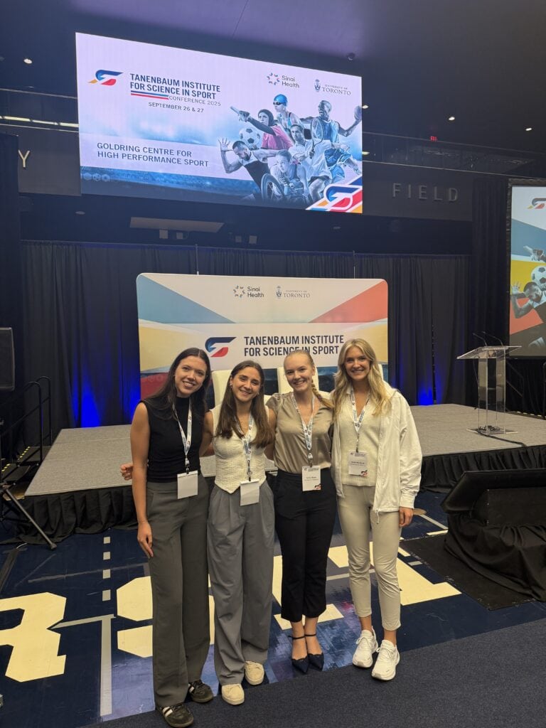 Four women at a sports science conference in front of a stage and presentation screens.